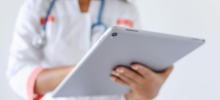 Une femme dont ne voit pas le visage mais on devine qu'elle porte un masque médical est en blouse blanche, avec un stéthoscope autours du coup et tien une tablette qui est en premier plan 