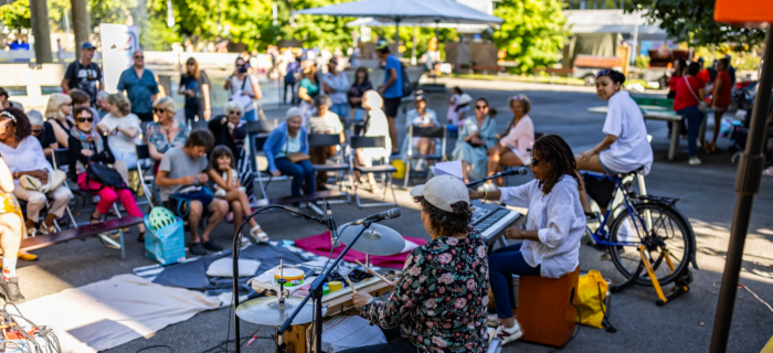Des personnes de diverses générations sont réunies en plein air et écoutent un groupe de musique