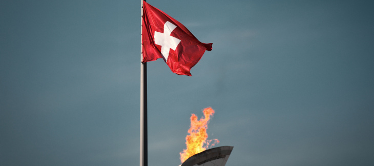 La flamme olympique brûle avec, sur sa gauche, le drapeau suisse flottant au vent